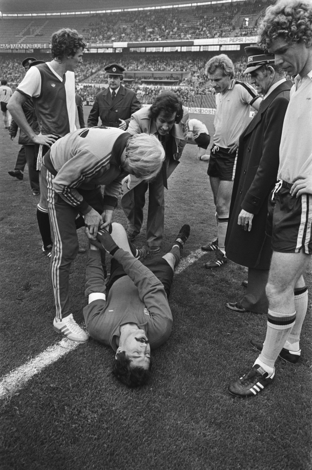 Schwarzes und weißes Bild eines Fußballspielers, der auf dem Boden liegt und von einer Gruppe Männer umgeben ist, mit einer Menge im Hintergrund des Stadions.