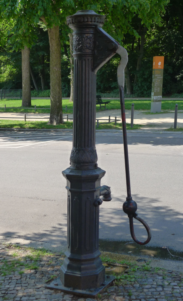 Eine alte Wasserpumpe im Park, umgeben von einer Straße, Gras, einer Bank, einer Platte und Bäumen.