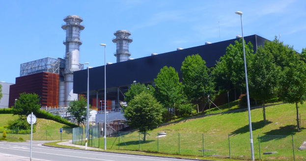 Großes Industriegebäude mit zahlreichen Fenstern, als Kraftwerk identifiziert, umgeben von Straßeninfrastruktur, Fahrzeugen, Grünflächen und einem bewölkten Himmel.