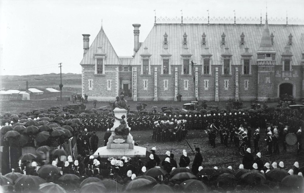 Eine große Menschenmenge steht vor einem Gebäude, einige halten Schirme, mit einer Statue auf einem Podest in der Mitte, Pfählen, Schuppen und einem klaren Himmel in einem Schwarz-Weiß-Foto der Einweihung der Königlichen kanadischen Mounted Police.