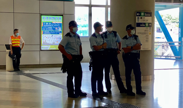 Eine Gruppe von maskierten Polizeibeamten steht vor einem Gebäude mit Glastüren, Schildern mit Text, einer Säule und einem Geländer, was darauf hinweist, dass sie Personen in Hong Kong festgenommen haben.