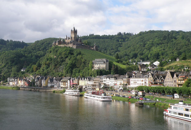 Eine malerische Aussicht auf den Rhein in Deutschland mit einer Burg auf einem Hügel, Booten auf dem Fluss und Fahrzeugen auf einer nahen Straße.