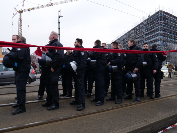Gruppe von Polizisten in schwarzen Uniformen und Helmen auf einem Bahngeleis mit einem roten Band davor, Fahrzeuge und Passanten im Hintergrund.