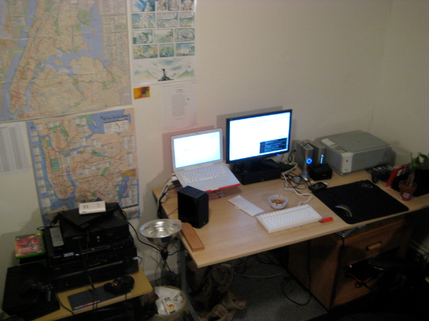 Ein Raum mit einem Tisch, auf dem ein Laptop, Lautsprecher, Monitor, Tastatur, Ball, Maus, Scanner und Papiere liegen, sowie ein weiterer Tisch mit Elektronikgeräten im Hintergrund, mit Charts und Postern an der Wand.