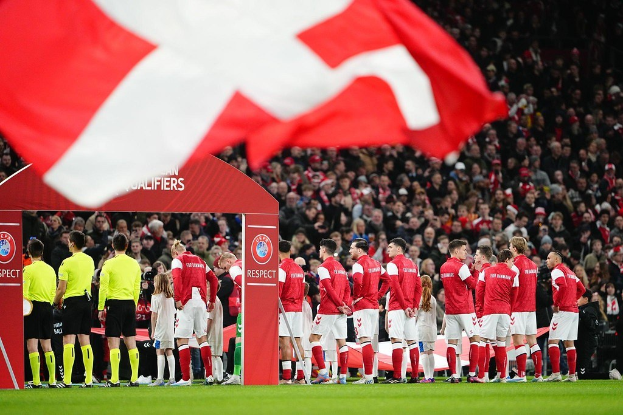 Eine Gruppe von Menschen auf einem Fußballfeld mit einer roten und weißen Flagge im Vordergrund, einem Bogen mit der Aufschrift "Bayern München vs Bayern Munich Betting Tips & Preview" im Hintergrund und einer großen Stadionmenge.