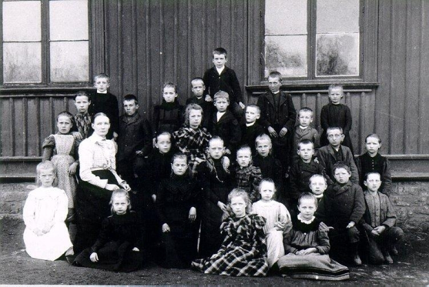 Ein Schwarz-Weiß-Foto einer Gruppe von Kindern, die vor einem Gebäude mit einer Wand und Fenstern im Hintergrund posieren, mit dem Text "1930er-Schulkinder" unten.