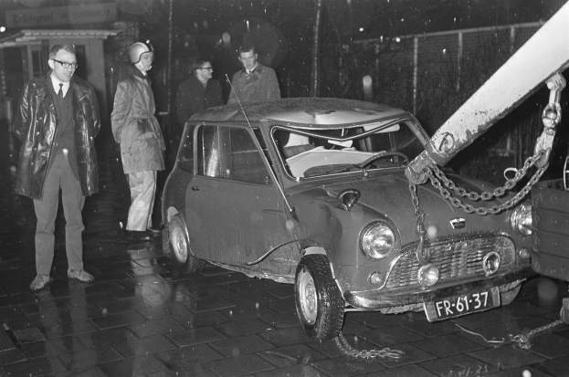 Ein Schwarz-Weiß-Bild eines Wagens, der von einem Abschleppwagen gezogen wird, umgeben von mehreren Menschen, einige tragen Helme, und einem Gebäude im Hintergrund.