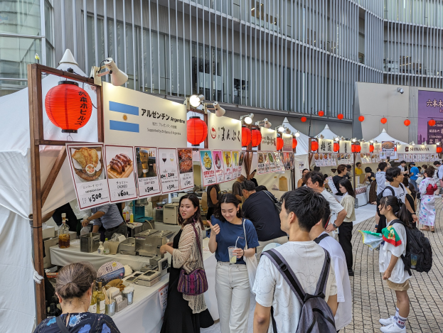 Menschen um einen tisch mit Essen auf einem Fest in Japan, mit Laternen und Informationsschildern im Hintergrund.