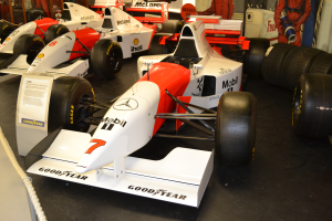 Eine Gruppe von Rennwagen in einem Museum ausgestellt, umgeben von Menschen, einem Textschild, einer Seilabsperrung und einer Wand im Hintergrund.