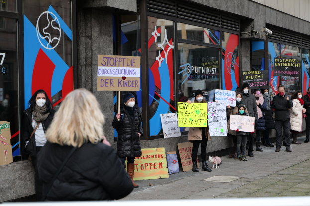 Eine Gruppe von Menschen mit Masken und Schildern protestiert vor einem Gebäude mit Glaswänden und -türen, einige mit Text und Logos auf Schildern, während Kameras und ein Hund anwesend sind.