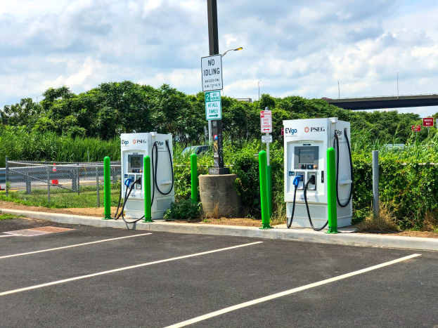 Elektrofahrzeug-Ladestation auf einem Parkplatz mit umgebenen Infrastruktur, Grünflächen und einer Brücke im Hintergrund.