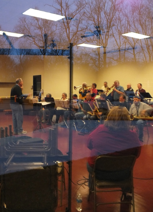 Musiker mit Trompeten im Hintergrund, Sänger vor Mikrofonen und eine Person, die Musik komponiert, mit einer Wand und Bäumen im Hintergrund durch eine Glasreflexion sichtbar