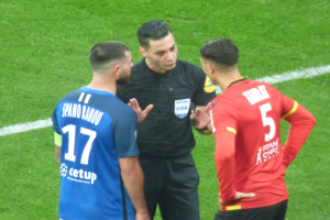 Schiedsrichter in schwarzem Hemd und Hose, der mit zwei Männern auf einem Fußballfeld spricht, einer in einem blauen und roten T-Shirt mit Text und Brille, der andere mit Brille.