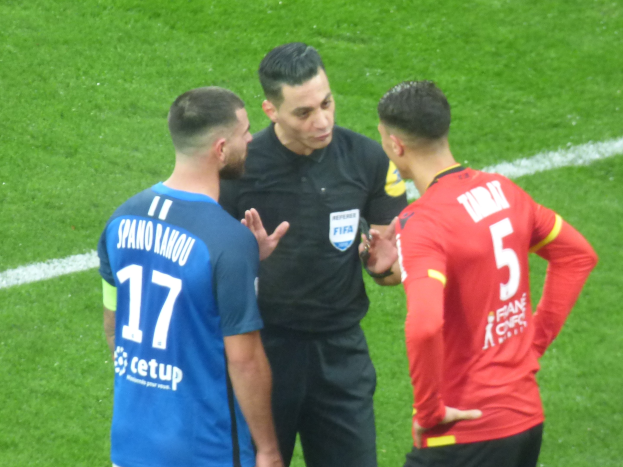 Schiedsrichter in schwarzem Hemd und Hose, der mit zwei Männern auf einem Fußballfeld spricht, einer in einem blauen und roten T-Shirt mit Text und Brille, der andere mit Brille.