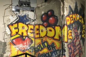 Mauer mit Graffiti, das in fetten schwarzen Buchstaben 'Freiheit und Demokratie' liest, ein Geländer unten und ein Gebäude mit Fenstern im Hintergrund.