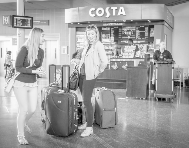Zwei Frauen mit Gepäck vor sich, mit einem Kaffeeladen im Hintergrund, wahrscheinlich am Flughafen.