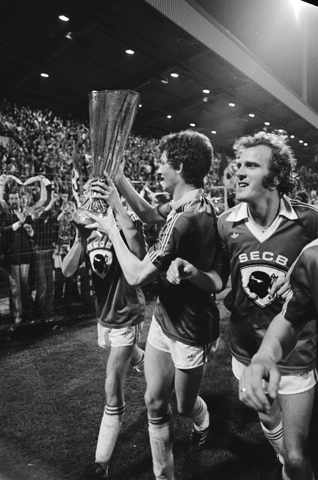 Schwarzes und weißes Bild von Fußballspielern, die mit einem Pokal auf dem Feld feiern, umgeben von einer Menge hinter einem Zaun unter Stadionbeleuchtung.