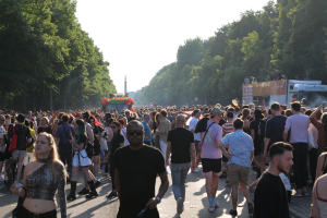 Eine große Menschenmenge, die eine von Bäumen gesäumte Straße entlanggeht, mit einem Turm im Hintergrund und Fahrzeugen mit Menschen auf der rechten Seite, wahrscheinlich während des Christopher Street Day in Berlin unter einem klaren blauen Himmel.