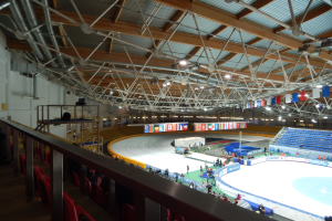 Große Indoor-Eisbahn mit zahlreichen Menschen, die Schlittschuh laufen, umgeben von Sitzplätzen, Geländern, Brettern, Beleuchtungskörpern und anderen Geräten, mit einer Wand im Hintergrund.