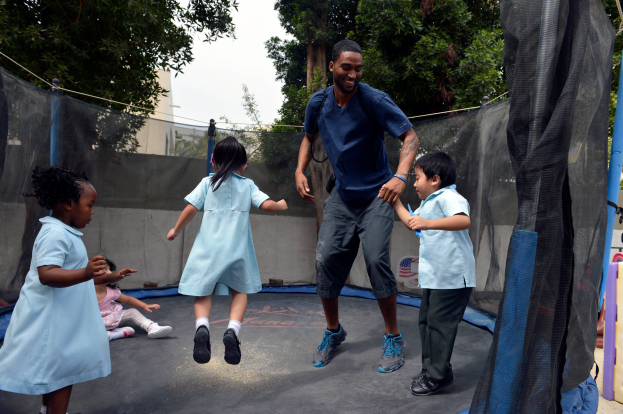 Eine Gruppe von Kindern, die auf einem Trampolin in einem Park spielen, umgeben von einem Netz, Stangen, einer Wand, Bäumen, Gebäuden und einem klaren blauen Himmel.