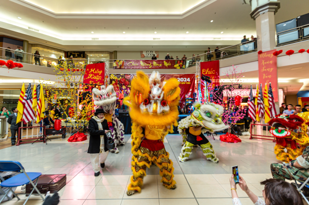 Ein lebendiges chinesisches Neujahrsfest in einem Einkaufszentrum mit Menschen in Kostümen, die tanzen, anderen, die auf Stühlen sitzen und Handys halten, umgeben von Flaggen, Bannern, Dekorationen, Geländern, Säulen und Deckenleuchten, mit einem Wasserzeichen unten.