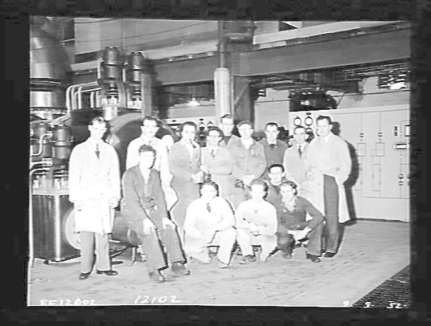 Eine Gruppe von Männern, die in einem 1940er-Jahre-Fabrik stehend und hockend, mit Maschinen und einer Wand im Hintergrund, schwarz-weiss.