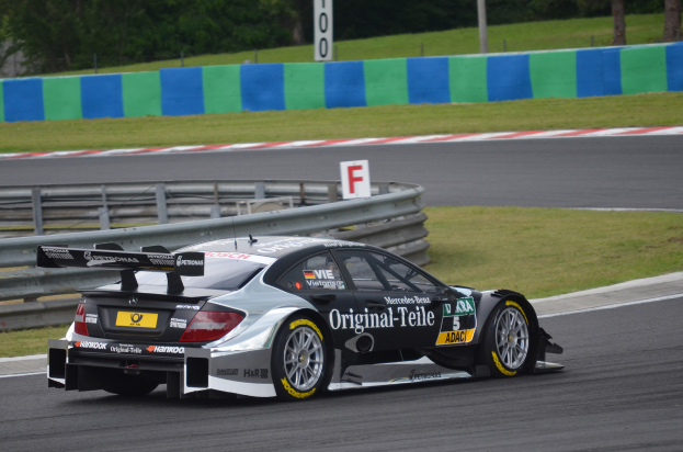 Ein Mercedes-Benz C63 AMG GT3 Rennwagen auf einer Rennstrecke, umgeben von Gras, einem Zaun, einer Tafel, Pfosten und Bäumen.
