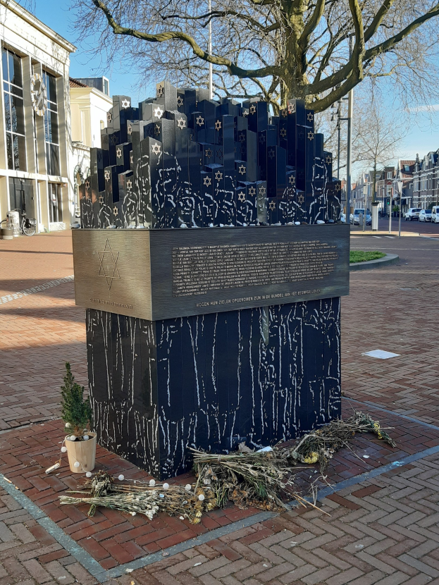 Denkmal in einem Stadtplatz für Holocaust-Opfer, mit einem Baum davor, einem Topf mit einer Pflanze, verstreuten trockenen Blättern und Gebäuden, Fahrzeugen, Pfählen und Bäumen im Hintergrund.