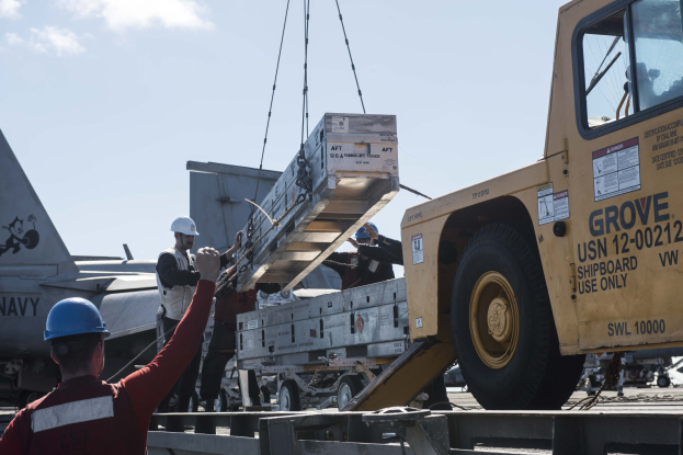 Ein Mann in rotem Hemd und blauer Helms hebt ein großes Gerät auf einen Lkw, mit einem Flugzeug und einem klaren blauen Himmel im Hintergrund.