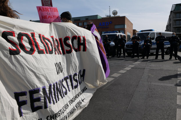 Eine Gruppe von Menschen marschiert die Straße entlang und hält ein Banner mit der Aufschrift "Solidarität und Feminismus" in der Hand, mit parkenden Fahrzeugen und Gebäuden im Hintergrund unter einem klaren blauen Himmel.