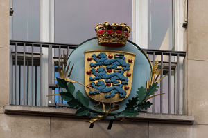 Wappen der britischen Königsfamilie an einem Gebäude mit einer Krone und einem Blatt-Logo, Fenstern und Metallgittern im Hintergrund.