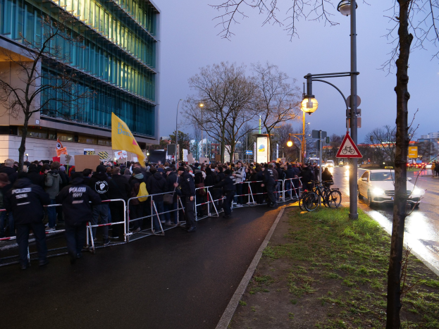Eine große Gruppe von Menschen demonstriert vor einem Gebäude in Berlin, hält Schilder, mit Barrikaden, Fahrrädern, Laternenpfählen, Schildern, Bäumen und Gras im Hintergrund.