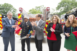 Gruppe von Menschen, die bei einer Ehrung der Alexandria Fire Department klatschen und lächeln, mit zwei Personen, die eine Schere und ein rotes Band halten, vor einem Feuerwehrauto.