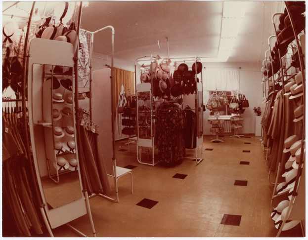Altes Schwarz-Weiß-Foto eines kleinen Bekleidungsgeschäfts-Innenraums mit Kleidungsstücken an Bügeln, Hüten auf Ständern, einem Spiegel, einem Stuhl, einem Tisch mit diversen Gegenständen, Vorhängen und einer Decke mit Einbauleuchten.