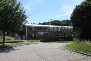 Ein Schulgebäude mit Fenstern, einem Laternenmast, einem Schild und einer Gruppe von Bäumen, umgeben von Gras und einem Zaun, unter einem bewölkten Himmel.