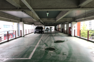 Ein mehrgeschossiges Parkhaus mit geparkten Autos, Gel├Ąndern, Pfeilern, Deckenleuchten, Rohren und umgebenden B├Ąumen, vor einer Kulisse von Geb├Ąuden mit Fenstern in einer Stadtlandschaft.