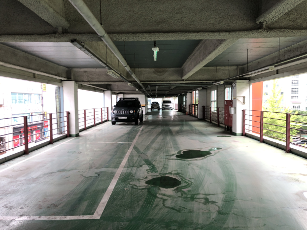 Ein mehrgeschossiges Parkhaus mit geparkten Autos, Gel├Ąndern, Pfeilern, Deckenleuchten, Rohren und umgebenden B├Ąumen, vor einer Kulisse von Geb├Ąuden mit Fenstern in einer Stadtlandschaft.