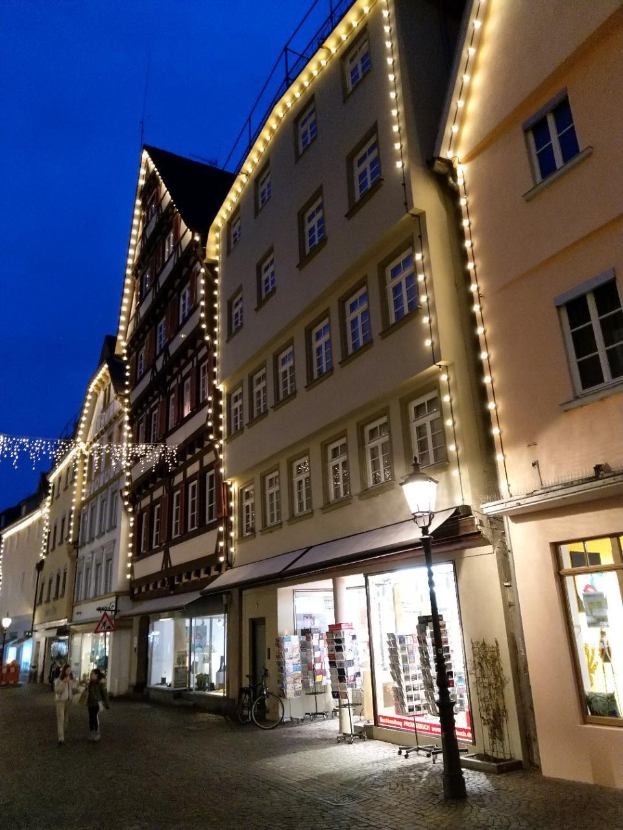 Eine festlich geschmückte Kopfsteinpflasterstraße in Rothenburg ob der Tauber, Deutschland, gesäumt von Häusern und mit Weihnachtslichtern geschmückt, wo Menschen spazieren gehen und ein Fahrrad geparkt ist.