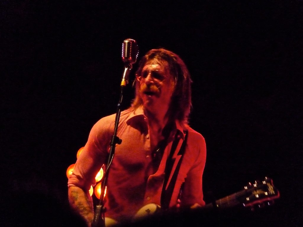 Eine Person mit Brille steht singend mit einer Gitarre in der Hand da und hat den Mund offen.