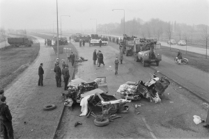 Schwarz-weiß-Szene eines Autounfalls am Straßenrand mit mehreren Fahrzeugen, darunter ein Landfahrzeug, und einer Gruppe von Menschen drumherum; Laternenpfähle, Bäume und Himmel im Hintergrund.
