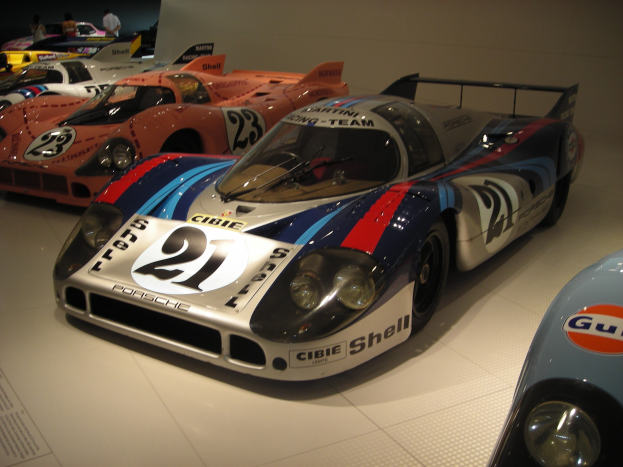 Porsche 917K Rennsportwagen in einem Museum neben anderen Sportwagen, mit Menschen und einer Wand im Hintergrund und informativem Text auf der linken Seite.