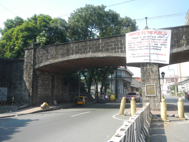 Eine Brücke mit einem "Hinweis an die Öffentlichkeit"-Schild, Fahrzeuge auf der Straße darunter, ein Geländer auf der rechten Seite und Gebäude, Bäume, Mäste, Drähte und der Himmel im Hintergrund.