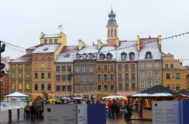 Ein lebendiger Weihnachtsmarkt im alten Stadtzentrum von Warschau mit Menschen um beleuchtete und geschmückte Stände, Gebäuden mit Fenstern im Hintergrund und einem Verkehrsampelmast auf der linken Seite.