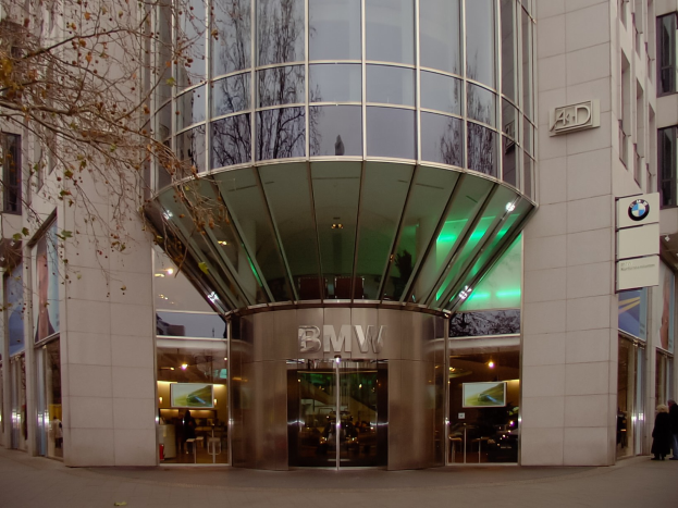 Moderner Glasfront-BMW-Hauptsitz in Frankfurt, Deutschland, mit Text an der Fassade, Menschen vor dem Gebäude und einem Baum links.