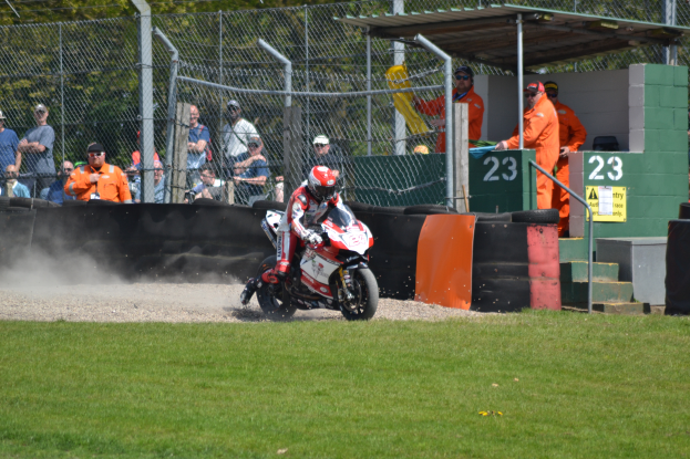 Eine Person auf einem Motorrad fährt über ein grasbewachsenes Feld, trägt einen Helm und Handschuhe, während Zuschauer mit Kappen und Schutzbrillen, ein Maschendrahtzaun, Pfähle, Bäume, Treppen mit Geländern und ein Schuppen mit Nummern und Texttafeln im Hintergrund zu sehen sind.