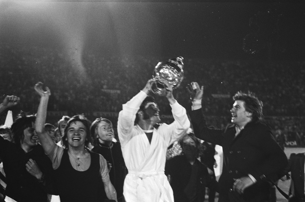 Eine Gruppe von Menschen, die mit einem Pokal feiern, wobei die zentrale Figur ihn triumphierend in die Höhe hält, vor einem jubelnden Publikum in Schwarz-Weiß.