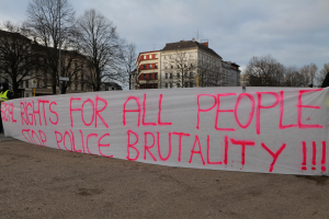 Eine Gruppe von Menschen, die auf dem Boden stehen und ein Banner halten, auf dem "Rechte für alle Menschen Stoppt Polizeigewalt" steht, mit einem Straßenschild, einem Schild, Bäumen, Gebäuden mit Fenstern und einem bewölkten Himmel im Hintergrund.