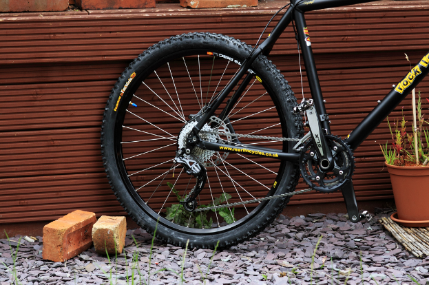Ein Fahrrad lehnt an einer Wand aus Ziegeln und Steinen, mit Pflanzen in der Nähe.