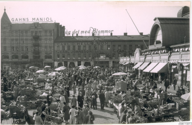 Schwarz-weiß-Foto eines belebten Berliner Marktes mit Menschen, Gemüsewagen und Gebäuden im Hintergrund.