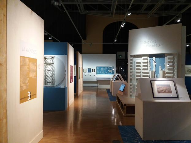 Ein Raum voller verschiedener Kunstwerke, darunter gerahmte Fotos auf Tischen, Texttafeln an den Wänden, Deckenleuchten und ein Teppichboden, was auf eine Ausstellung im Museum of Modern Art hindeutet.